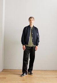 Man standing against plain white wall wearing black bomber jacket, olive green shirt, black pants, and black sneakers with white stripes on wooden floor.