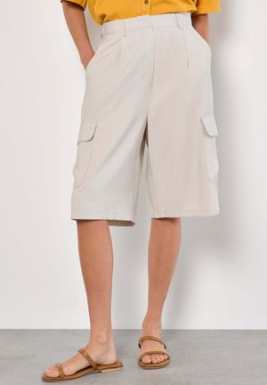 Shorts cargo beiges en tissu léger, dotés de deux poches latérales et d'une coupe décontractée, se terminant au-dessus du genou. Portés avec des sandales brunes.