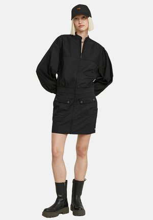 Schwarzes Zip-Jackenkleid mit hohem Kragen, oversized Ärmeln und Fronttaschen. Kombiniert mit knielangen schwarzen Stiefeln und einer schwarzen Mütze.