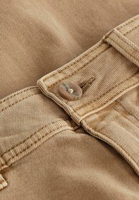 Beigenvärinen puuvillakangas, jossa on kevyt jousto. Siinä on ommeltu vyötärönauha ja pyöreä ruskea nappi, jossa on teksturoitu pinta.