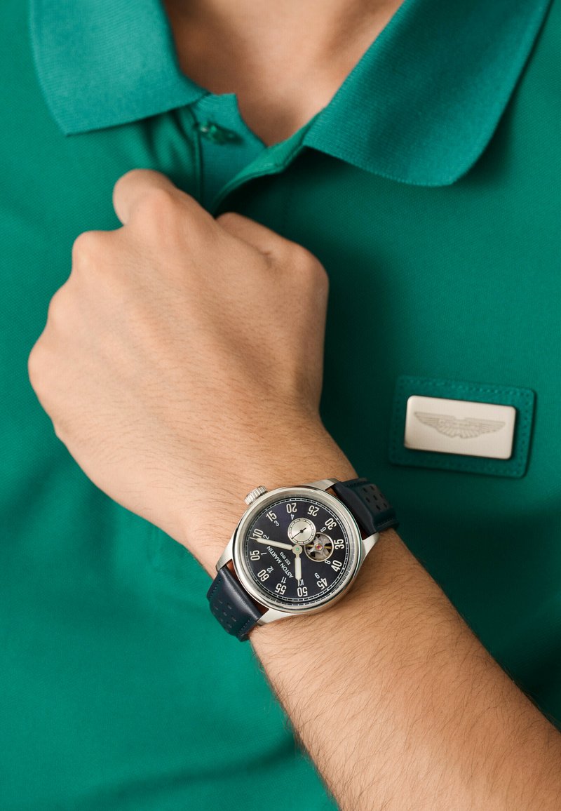 Reloj plateado con correa de color azul oscuro, cara redonda y esfera negra que muestra marcadores de hora blancos. Camiseta polo verde con un parche de marca.