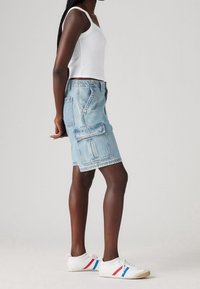 Mujer vestida con una camiseta sin mangas blanca, una falda cargo de denim azul claro y zapatillas blancas con rayas rojas y azules, de pie de lado.