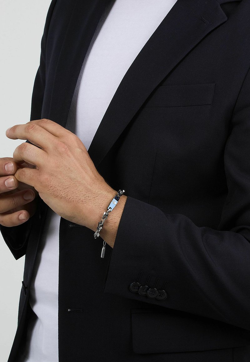 Bracciale a catena in tono argento con charm, caratterizzato da una combinazione di maglie piatte e texture. Indossato su un polso con un blazer blu navy e una camicia bianca.