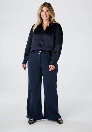 Lachende vrouw met lang blond haar, die een donkere zijden blouse, wijde marineblauwe broek met riem en zwarte studded ballerina's draagt, staande tegen een witte achtergrond.