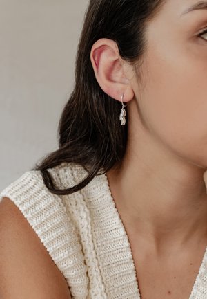 Silberne Tropfohrringe mit einem strukturierten Design, die an einem dünnen Ring hängen. Getragen von einer Person mit langen, dunklen Haaren, vor einem neutralen Hintergrund.