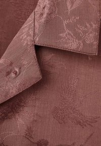 Struktureret mauve stof med et intrikat mønster af fugle og løv; har en krave og en glat overflade.