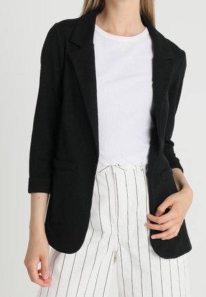 Femme portant un blazer noir aux manches retroussées, un haut blanc côtelé à manches courtes et un pantalon blanc taille haute avec des rayures noires verticales.
