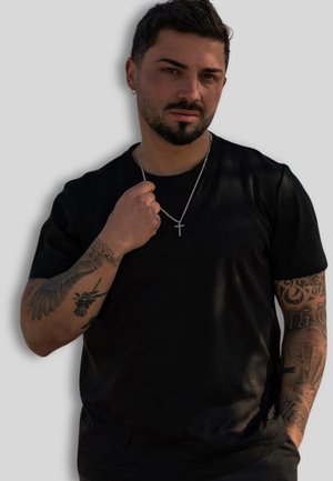 Schwarzes Baumwoll-T-Shirt mit kurzen Ärmeln und rundem Halsausschnitt. Silberne Kreuzkette, filigrane Tattoo-Designs auf den Armen sichtbar. Neutraler Hintergrund.