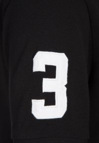 Écusson blanc numéro 3 brodé sur le tissu noir d’une manche de vêtement.