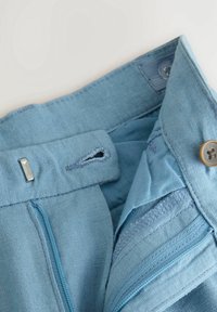 Gros plan sur une ceinture en lin bleu clair avec un bouton, une fermeture éclair, une boutonnière et un détail de crochet ajustable.
