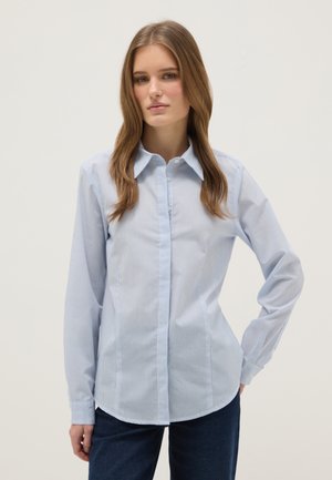 Joven con cabello largo castaño claro, lleva una camisa de botones a rayas azul claro y pantalones oscuros, de pie con una mano en la cadera.