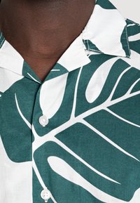 Camicia a maniche corte con bottoni, verde e bianca, con grandi motivi di foglie, realizzata in un tessuto liscio. Presenta un colletto classico e bottoni.