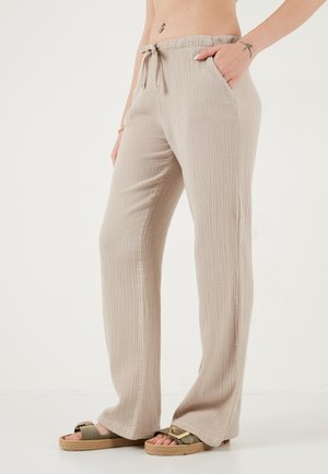 Pantalon large beige texturé avec taille à cordon, porté par une personne ayant une main dans la poche et des sandales à boucles dorées.