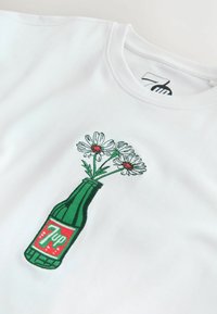 Chemise blanche avec une bouteille verte de 7up brodée tenant trois marguerites blanches aux centres rouges sur le centre de la poitrine devant.