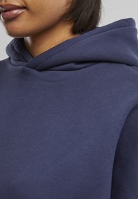 Sweat à capuche bleu marine avec une texture douce, doté d'une capuche à cordon de serrage et d'une coupe décontractée ; coutures visibles et surface du tissu lisse.