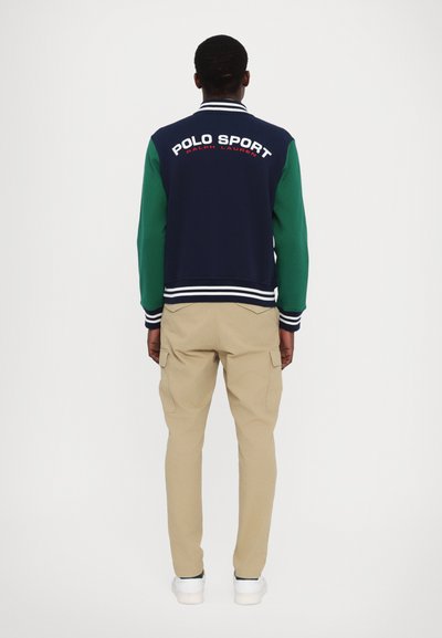 Marineblå og grønn bomberjakke med hvite striper, med "POLO SPORT" logo på baksiden. Kombinert med beige bukser og hvite sko.