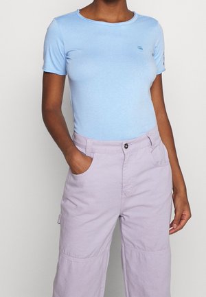 Persoon draagt een lichtblauw aansluitend T-shirt met korte mouwen, ingestopt in een lichtpaarse broek met hoge taille, handen in de zakken.