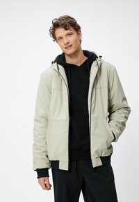 Lichtgrijze, capuchonjack met contrasterende zwarte accenten en een gladde, water resistente stof. Beschikt over een ritssluiting aan de voorkant en zakken.