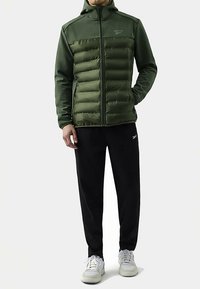 Dunkelgrüne Steppjacke mit Reißverschluss vorne, strukturierten Einsätzen und Kapuze, kombiniert mit schwarzer Hose und weißen Sneakers.