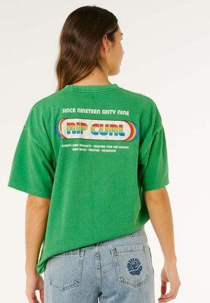 Frau mit langen braunen Haaren trägt ein grünes Rip Curl T-Shirt mit Regenbogenlogo und hellblaue Jeans mit blauer Blumenstickerei auf der Tasche.