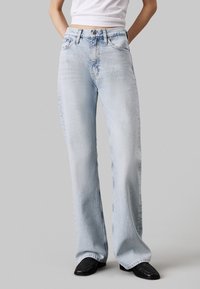 Vaqueros de mezclilla de pernera ancha en azul claro, con cintura alta, diseño clásico de cinco bolsillos y un ligero desgaste en las rodillas y los muslos.
