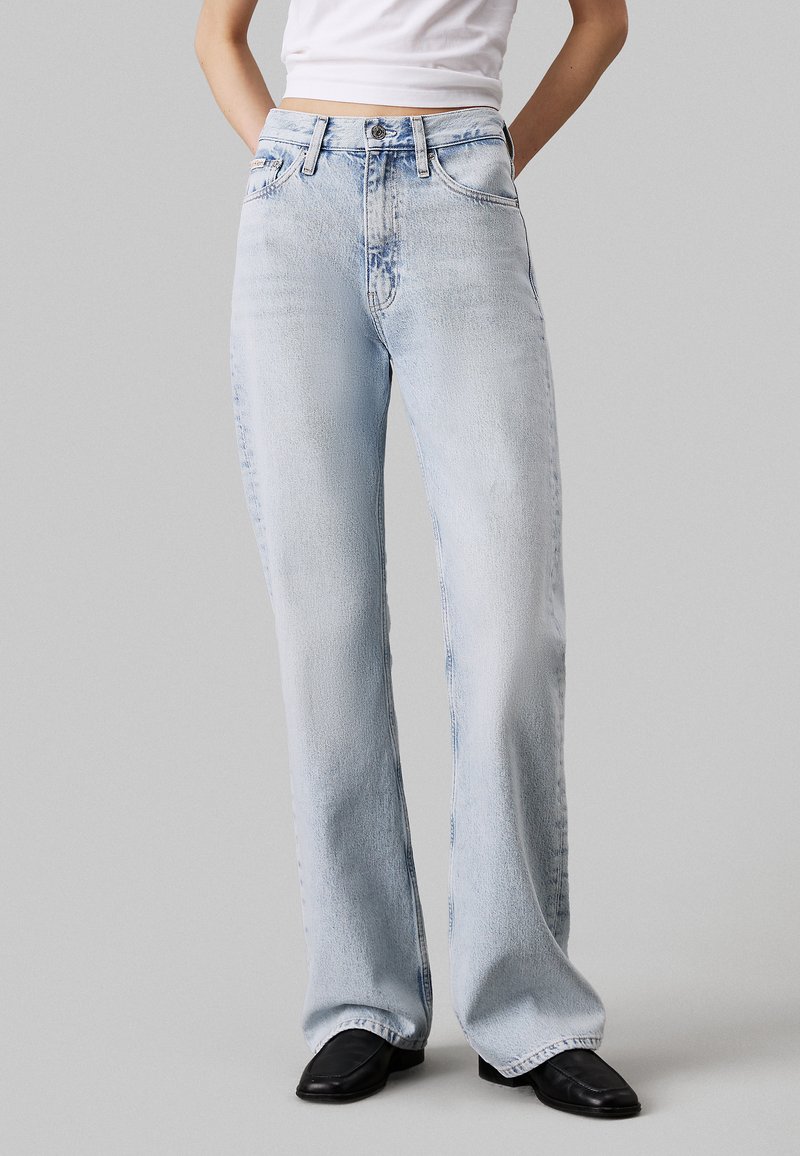 Vaqueros de mezclilla de pernera ancha en azul claro, con cintura alta, diseño clásico de cinco bolsillos y un ligero desgaste en las rodillas y los muslos.