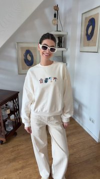 Χαμογελαστή γυναίκα που φοράει λευκά γυαλιά ηλίου, κρεμ φούτερ με πολύχρωμα φυτικά σχέδια και κρεμ παντελόνι με ρίγες σε ένα δωμάτιο με ξύλινο πάτωμα.