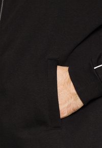 Sweat-shirt noir avec une fermeture éclair, texture douce et une poche latérale avec une main visible insérée. Design simple et minimaliste.