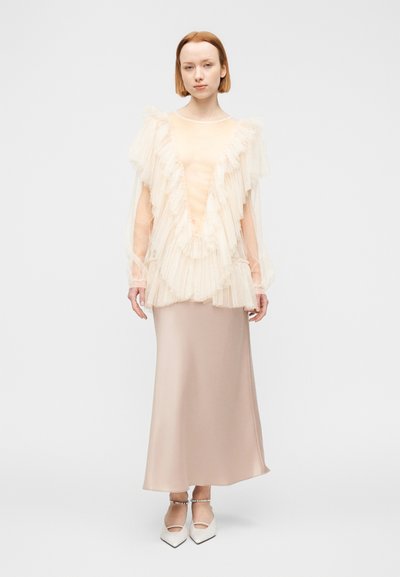 Ανοιχτόχρωμο τοπ με βολάν και διαφανή στρωματοποιημένα μανίκια · εφαρμοστή σατέν φούστα σε γυμνό χρώμα · λευκά παπούτσια με τακούνι και λουρί γύρω από τον αστράγαλο. Ομαλές υφές σε όλη την εμφάνιση.