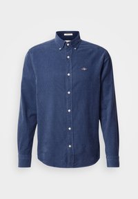 Chemise en velours côtelé bleu foncé à manches longues avec col boutonné, boutons blancs et petit logo brodé sur la poitrine gauche.