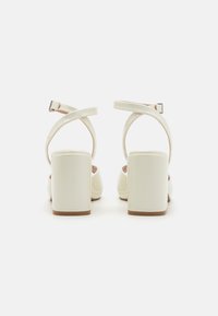 Weiße Sandalen mit Absatz, Blockabsatz, gekreuzten Knöchelriemen, glatter Oberfläche und minimalistischem Design. Weiche Innenauskleidung sichtbar.