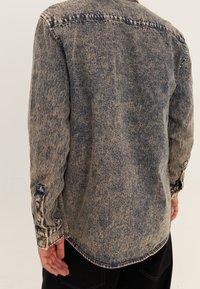 Koszula jeansowa w wyblakłym niebieskim odcieniu, z długimi rękawami, mankietami na guziki i zaokrąglonym dołem. Postarzany materiał tworzy vintage'owy wygląd.