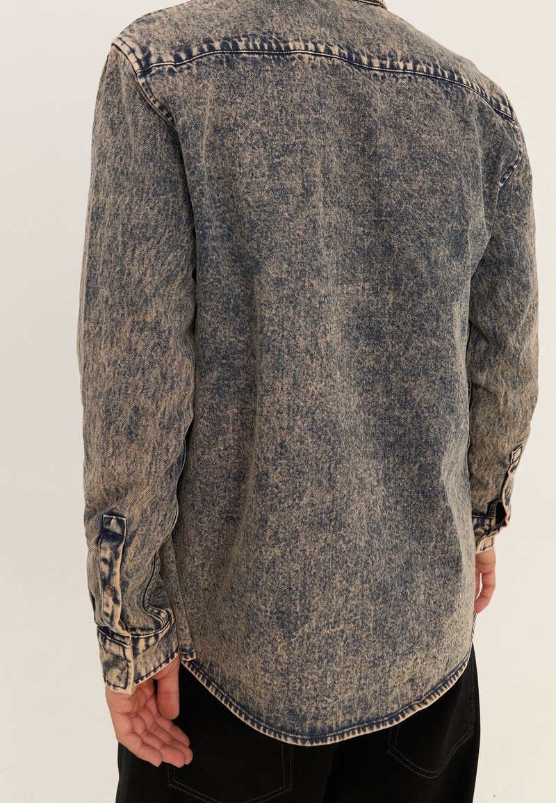 Koszula jeansowa w wyblakłym niebieskim odcieniu, z długimi rękawami, mankietami na guziki i zaokrąglonym dołem. Postarzany materiał tworzy vintage'owy wygląd.