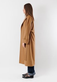 Cappotto lungo color cammello realizzato in tessuto testurizzato con una cintura in vita. Abbinato a jeans scuri e ballerine nere arrotondate.