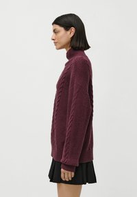 Kvinne i profil som har på seg en bordeaux kabelstrikket turtleneck-genser og et svart plisségjør mot en enkel hvit bakgrunn.