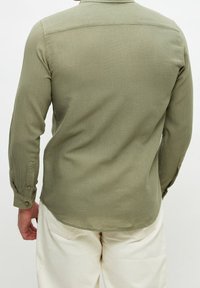 Camisa de manga larga verde oliva con una tela texturizada, puños abotonados y un dobladillo curvado. La vista trasera muestra un diseño limpio y sencillo.