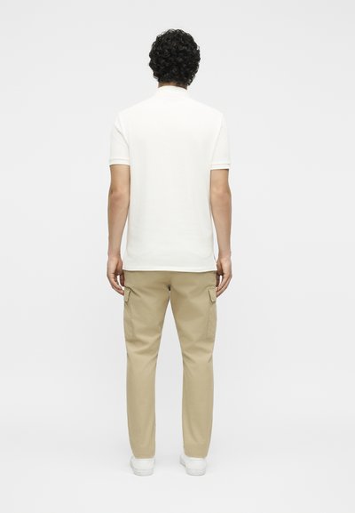 Άνδρας που στέκεται με την πλάτη προς τον θεατή, φοράει λευκό κοντομάνικο πουκάμισο, μπεζ παντελόνι cargo και λευκά αθλητικά παπούτσια σε μονόχρωμο φόντο.