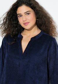 Nainen, jolla on kihara tumma hius, käyttää kultaisia renkaankorviksia ja tummansinistä struktuuripintaista pitkähihaista paitaa, hymyilee lempeästi valkoista taustaa vasten.