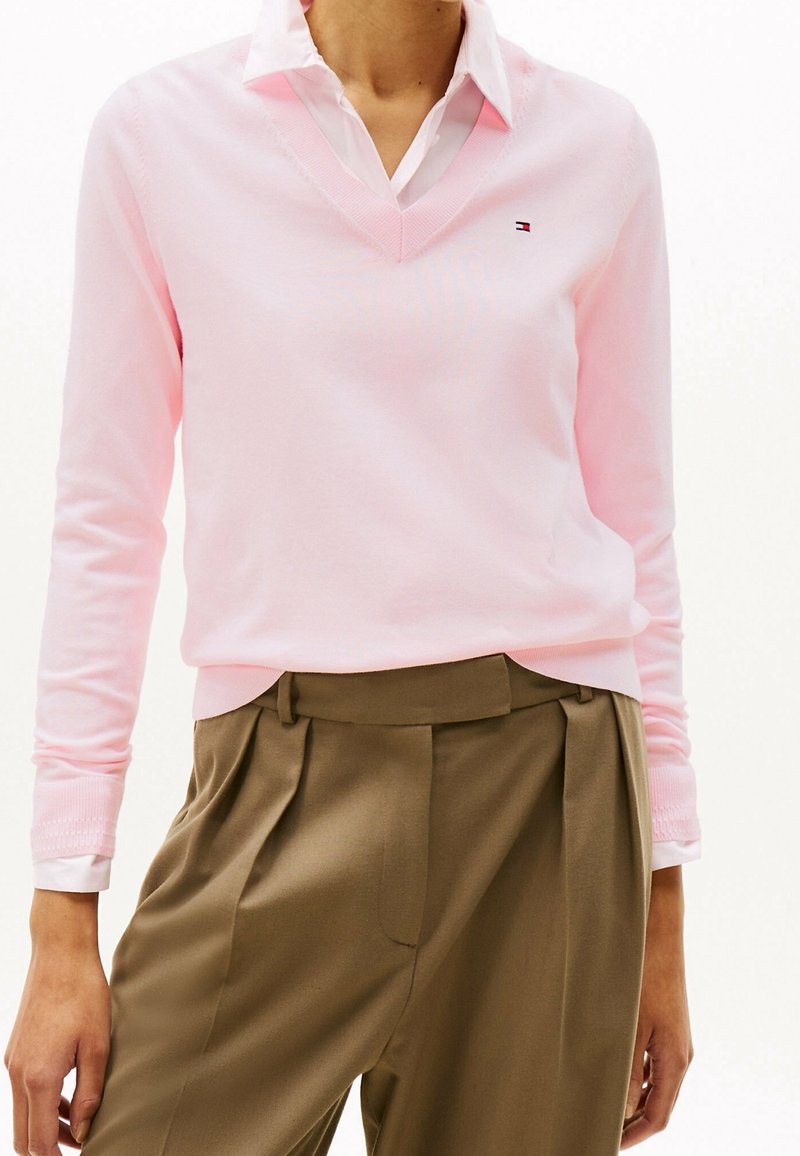 Maglione rosa chiaro a scollo a V in morbido tessuto, con maniche lunghe e polsini a coste. Indossato sopra una camicia bianca con colletto e pantaloni beige.