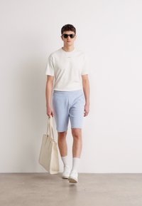 Lagani bijeli T-shirt, svijetloplave kratke hlače, bijele tenisice i bež torba. Model nosi tamne sunčane naočale, stoji na neutralnom podu.