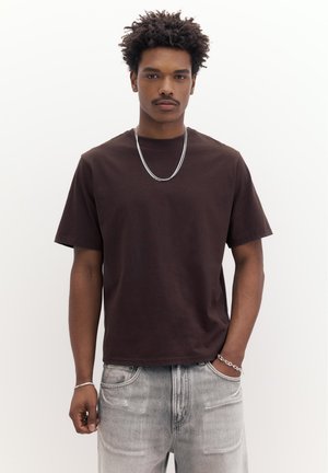 Mladenič z kodrastimi lasmi, oblečen v rjavo majico s kratkimi rokavi, srebrno verižico, sive kavbojke in srebrno zapestnico, ki stoji pred belim ozadjem.