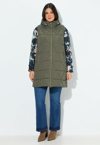 Zelená prošívaná vesta s kapucí, na zip, a bočními kapsami, nošená přes vzorovaný top s dlouhým rukávem, v kombinaci s modrými rozšířenými džíny.