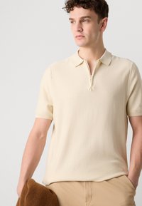 Beige, kortärmad polo med dragkedja i kragen, tillverkad av strukturerad stickad material. Bärs med bruna byxor och håller en brun tröja.