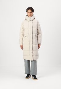 Manteau d'hiver long, de couleur claire, avec un col haut, un motif matelassé, des zips sur les côtés et deux poches avant, associé à un jean large et des baskets.