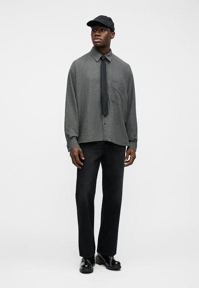 Άνδρας που φοράει γκρι πουκάμισο με μοτίβο και μακριά μανίκια, μαύρη γραβάτα, μαύρο παντελόνι, μαύρο καπέλο και γυαλιστερά μαύρα μποτάκια, στέκεται μπροστά σε λευκό φόντο.