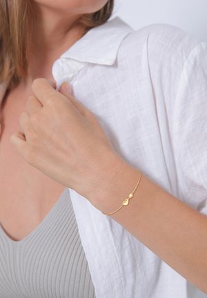 Bracciale d'oro con un charm a forma di cuore e stella, indossato su una mano dalla pelle chiara. Sullo sfondo si vede un morbido tessuto bianco.