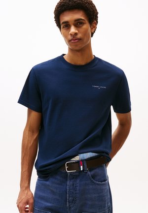 Ung mand iført en mørkeblå Tommy Jeans T-shirt, blå jeans og et sort bælte med en rød, hvid og blå detalje på spændet.