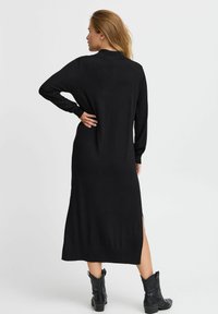 Vestido negro de manga larga con aberturas laterales, puños acanalados y dobladillo. La suave tela cae hasta el tobillo, complementado con botas negras por el tobillo.