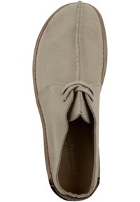 Beige mockaskosko med rund tå, central söm och snörning. Insidan har en kontrasterande brun innersula och en texturerad sula.