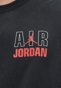 Μαύρο ύφασμα με λευκά γράμματα περιγράμματος «AIR», μία κόκκινη σιλουέτα καλαθοσφαιριστή που πηδά στο κέντρο και έντονο κόκκινο κείμενο «JORDAN» από κάτω.